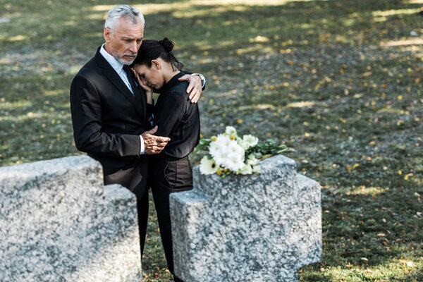 селективное внимание бородатого мужчины в костюме, обнимающего привлекательную женщину и гробницы
 