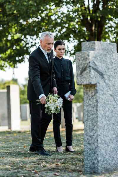 selective focus of bearded man in suit holding flowers near attractive woman in graveyard 