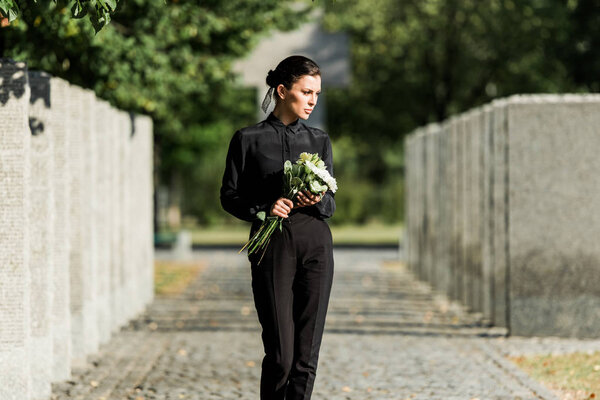 sad woman holding white flowers and walking in graveyard 