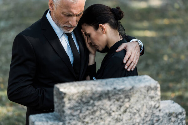 selective focus of senior man hugging upset woman near tombstone 