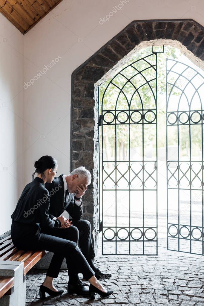 beautiful woman sitting on bench near upset senior man in cemetery 