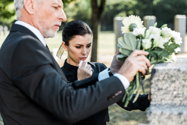 selective focus of senior man holding flowers near tomb and woman 