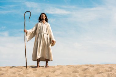 handsome man in jesus robe holding wooden cane against blue sky in desert  clipart