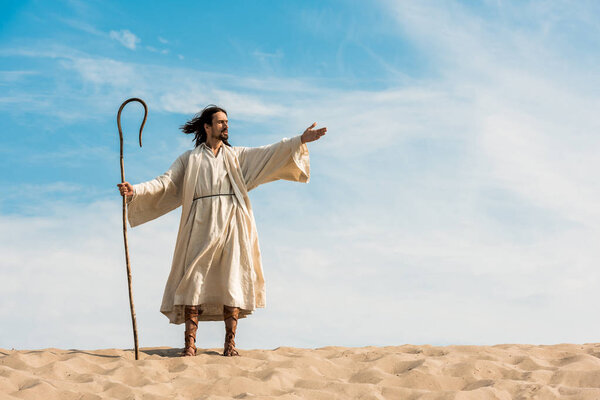 man in jesus robe holding wooden cane and gesturing against blue sky in desert 