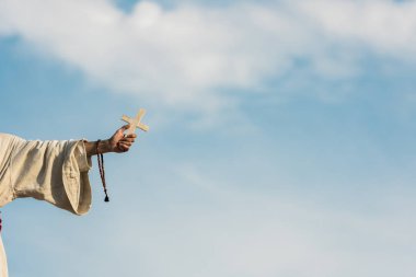 cropped view of religious man holding cross and rosary beads against blue sky  clipart