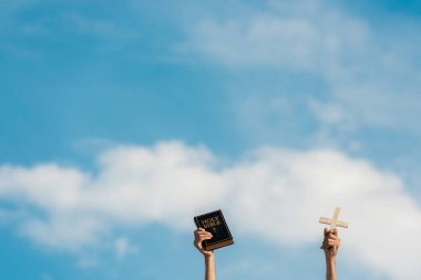 cropped view of man holding holy bible and cross against blue sky with clouds clipart