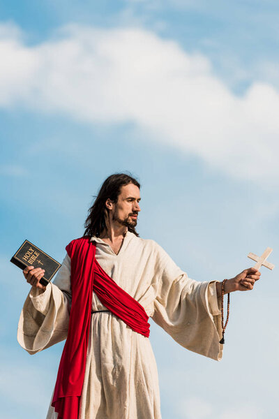Иисус держит священный библейский и деревянный крест против голубого неба с облаками
 