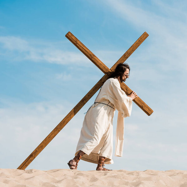 bearded man walking with wooden cross against blue sky in desert 