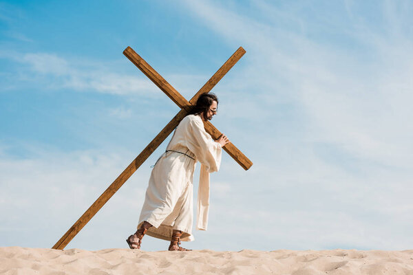 bearded man walking with wooden cross against sky in desert 