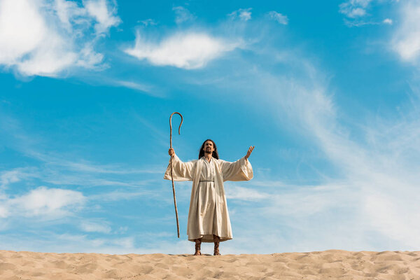 jesus with outstretched hands holding wooden cane against blue sky in desert 