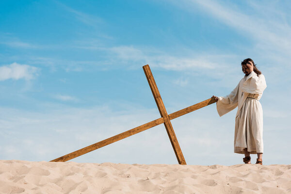 bearded man standing near wooden cross in desert on sand 