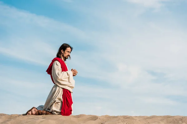 jesus praying on knees with clenched hands in desert against blue sky