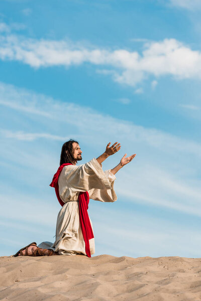 man praying on knees with outstretched hands in desert against blue sky 