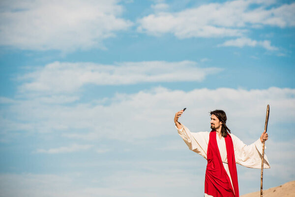 handsome man in jesus robe talking selfie while holding wooden cane 