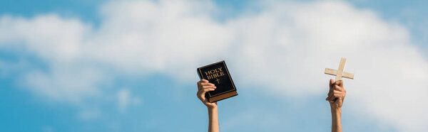панорамный снимок человека, держащего святую Библию и крест на голубом небе с облаками
