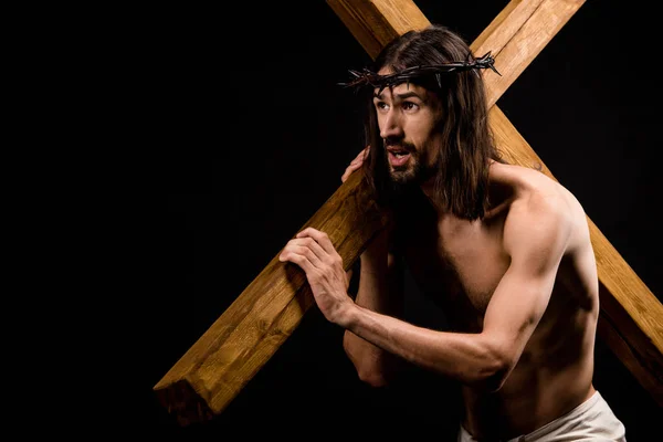 黒で隔離された重い木製の十字架を保持する花輪のシャツなしのイエス — ストック写真