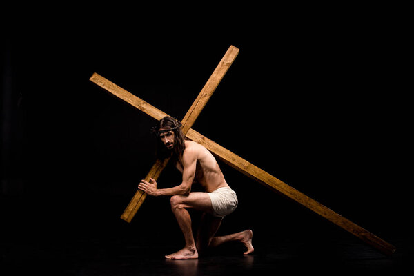 shirtless jesus sitting and holding wooden cross on black 