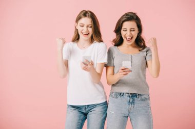 çekici ve gülümseyen kadınlar t-shirt akıllı telefonlar tutan ve evet jest gösteren pembe izole