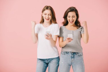 attractive and smiling women in t-shirts holding smartphones and showing yes gesture isolated on pink clipart