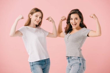 attractive and smiling women in t-shirts showing muscles and looking at camera isolated on pink clipart