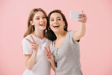 attractive and smiling women in t-shirts taking selfie and showing peace signs isolated on pink clipart