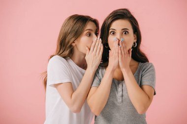 attractive woman telling secret to her shocked friend in t-shirt isolated on pink  clipart