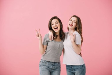 attractive and smiling women in t-shirts looking at camera and showing peace sign isolated on pink clipart