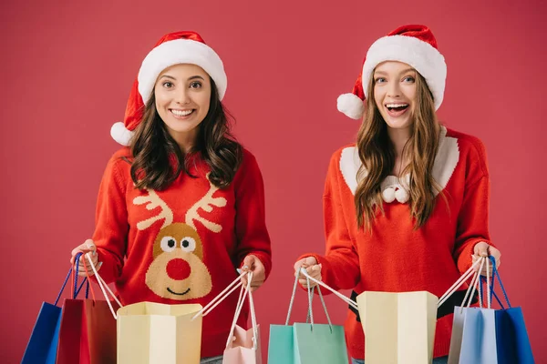 Aantrekkelijke Lachende Vrouwen Truien Santa Hoeden Houden Boodschappentassen Geïsoleerd Rood — Stockfoto