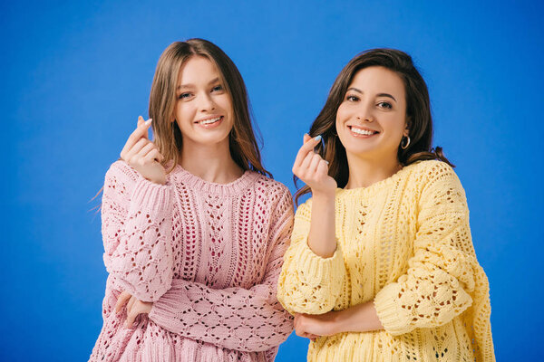 attractive and smiling women in sweaters showing heart gesture isolated on blue 