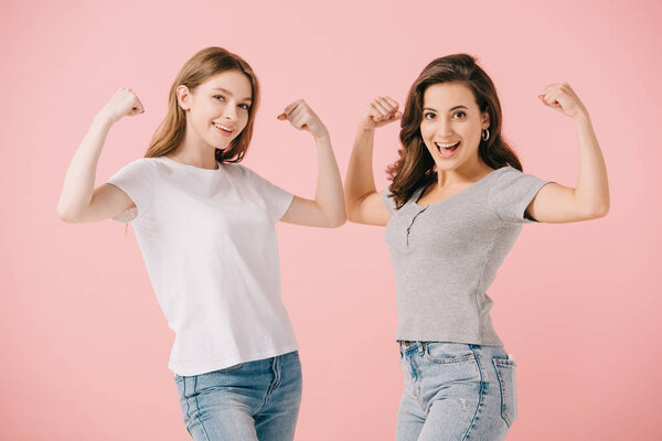 attractive and smiling women in t-shirts showing muscles and looking at camera isolated on pink