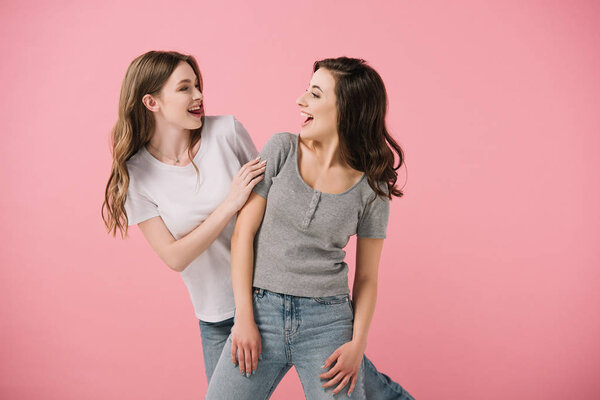 привлекательные и улыбающиеся женщины в футболках, смотрящие друг на друга изолированно на розовом
