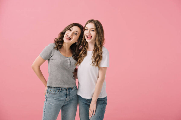 привлекательные и улыбающиеся женщины в футболках с торчащими языками, смотрящие в камеру, изолированную на розовом
