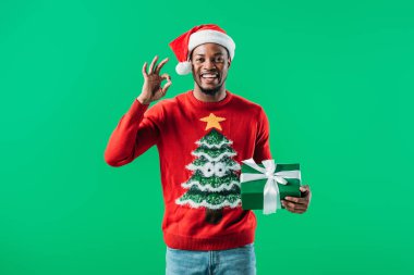 Noel kazak ve Santa şapka Afro-Amerikan adam tamam işareti gösteren ve yeşil izole hediye kutusu tutarak