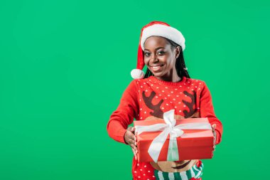 Noel kazak ve Santa şapka beyaz kurdele ile hediye kutusu tutan ve yeşil izole kamera bakarak Afrikalı Amerikalı kadın