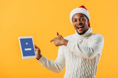 Kiev, Ukrayna - 2 Ağustos 2019: Noel Baba şapkalı Afro-Amerikan adam, Facebook uygulaması sarı yalıtılmış dijital tablette parmakla işaret ediyor