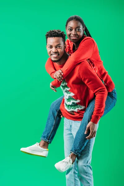 African American man holding on back woman in Christmas sweater isolated on green
