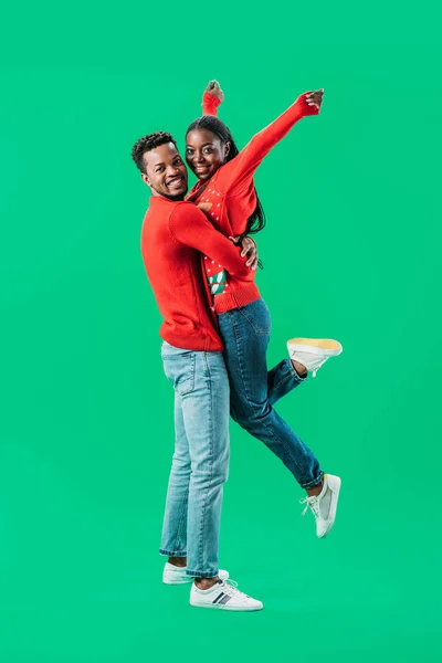 赤いクリスマスセーターを着たアフリカ系アメリカ人男性が 緑色に孤立した手を挙げた女性を抱きしめる — ストック写真