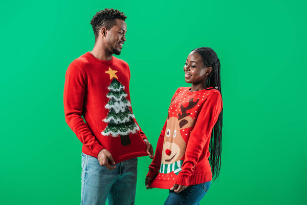 African American man near woman in red Christmas sweaters smiling and looking at each other isolated on green