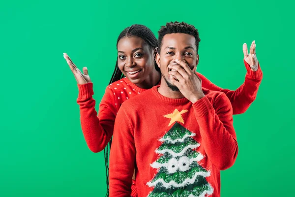 African American woman raising hands behind surprised man in Christmas sweater isolated on green