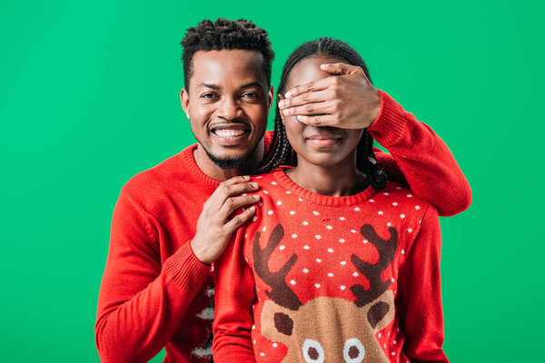 African American man in red sweater covering eyes with hand to woman and looking at camera isolated on green