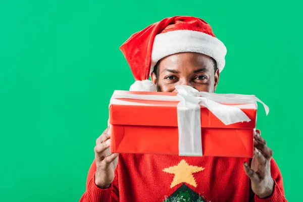 在圣诞老人帽子的非洲裔美国人拿着红色的礼物盒与白色丝带 看着相机隔离在绿色 — 图库照片