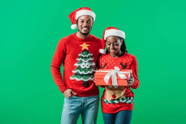 非洲裔美国男子在红色圣诞毛衣和圣诞老人帽子附近妇女拿着礼物盒孤立在手中的绿色 — 图库照片