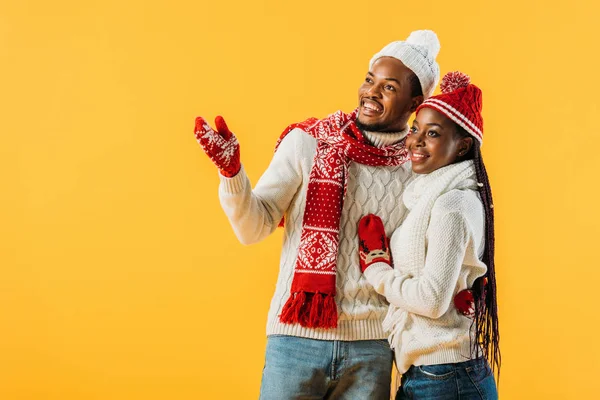 African American man in winter outfit hugging woman and pointing with hand isolated on yellow