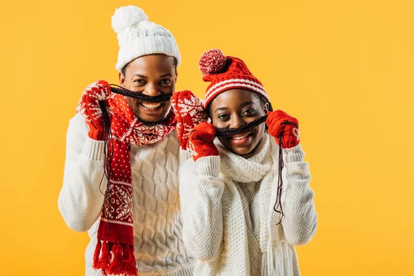 African American couple in winter outfit making mustache with hair and looking at camera isolated on yellow