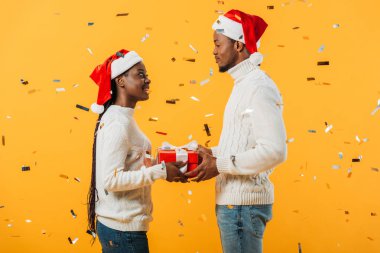 side view of African American couple in Santa hats holding gift box and looking at each other on yellow background with confetti