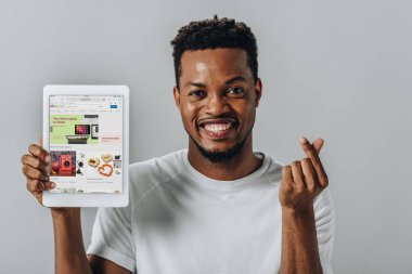 KYIV, UKRAINE - AUGUST 2, 2019: African American man holding digital tablet with ebay website and showing money gesture isolated on grey