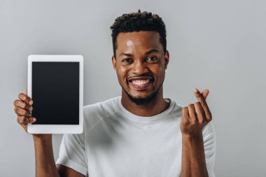 African American man holding digital tablet with blank screen and showing money gesture isolated on grey