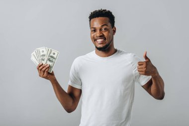 African American man holding dollar banknotes and showing thumb up isolated on grey
