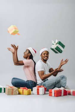 Santa şapkalı Afro-Amerikan çift arka arkaya oturan ve gri izole hediye kutuları kusma