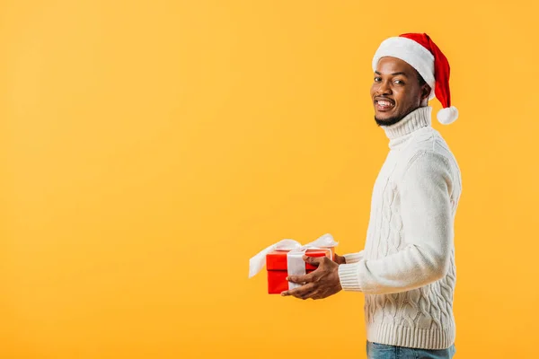 侧视图的非裔美国人在圣诞老人帽子拿着礼品盒 看着相机隔离在黄色 — 图库照片